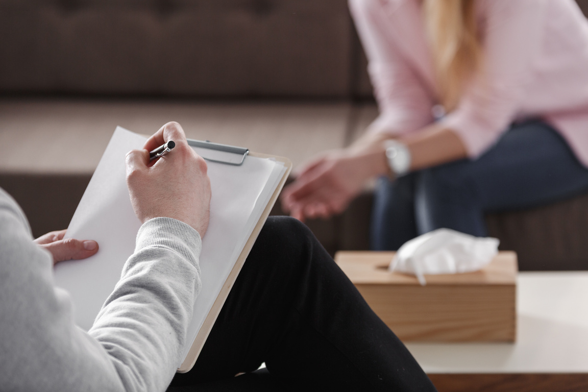 Close-up of therapist hand writing notes during a counseling session with a single woman sitting on a couch in the blurred background.